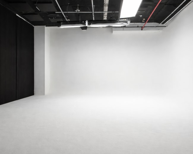 The image shows an empty, minimalist studio space with white walls and a smooth floor, illuminated by overhead lights.