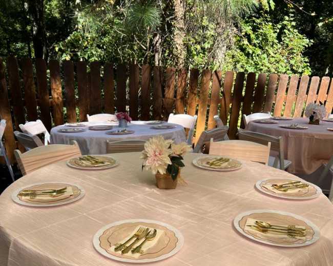 A round table is elegantly set for a meal outdoors, featuring gold utensils and pink tablecloths, surrounded by white chairs.