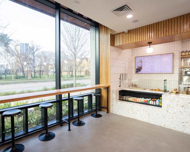 The image shows a modern café interior with a countertop, menu board, and several black stools facing large windows that overlook a park area.