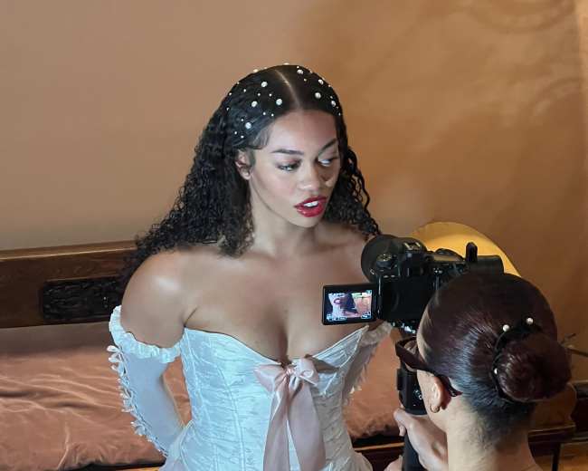 A woman in a white corset and dress is being filmed by another person holding a camera in a dimly lit room.