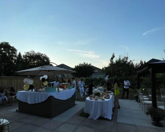 A backyard party features tables adorned with food and drinks, surrounded by guests enjoying the evening under a clear sky.