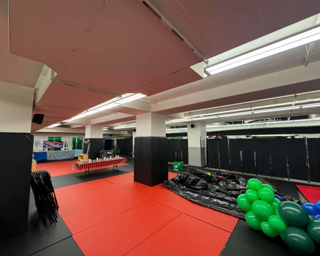 The image shows a spacious indoor area with red and black padded flooring, surrounded by black padded walls, and features tables and equipment on display.