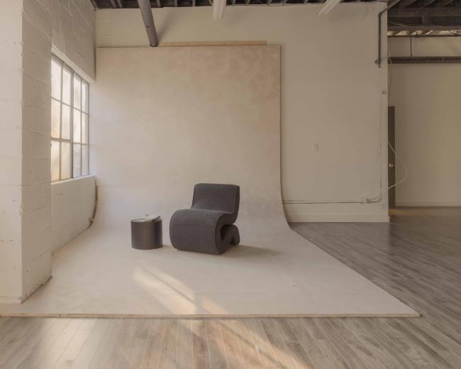 The image shows a minimalist studio space with a single chair and a small table on a light-colored backdrop, illuminated by natural light through large windows.