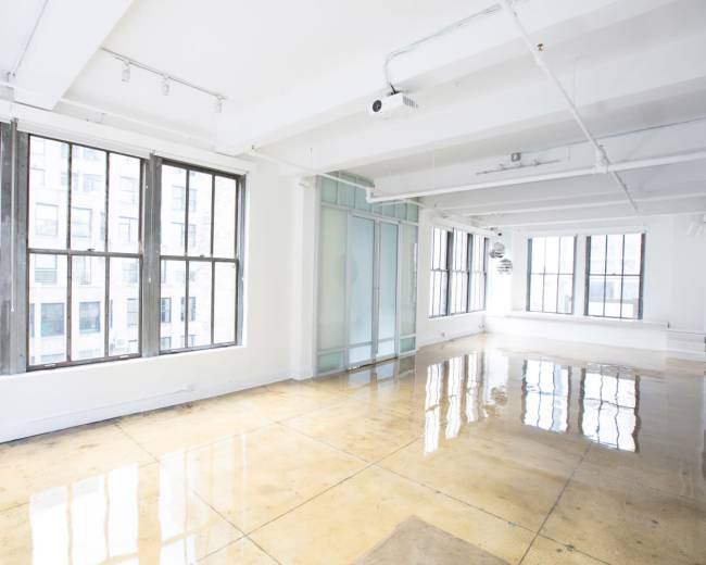 The image shows a spacious, bright room with large windows, polished concrete floors, and an open layout.