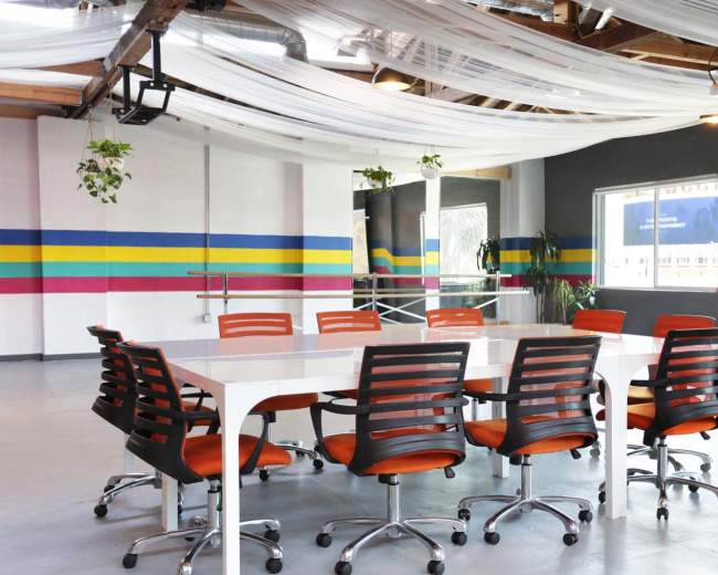 The image shows a modern conference room with a large white table surrounded by orange and black chairs, set against colorful striped walls.