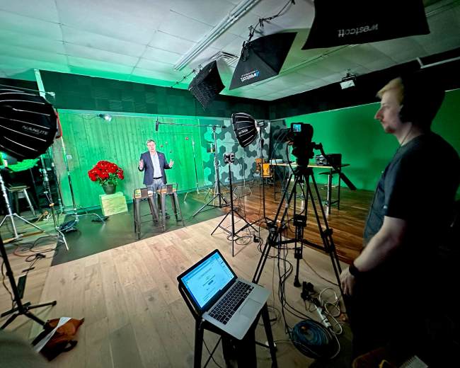 A man stands on a set with green screens and studio lights, while a crew member operates a camera and monitors equipment.