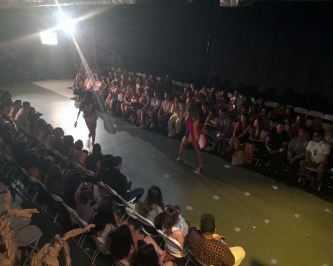 A fashion runway show features models walking down the catwalk while an audience watches from seated rows.