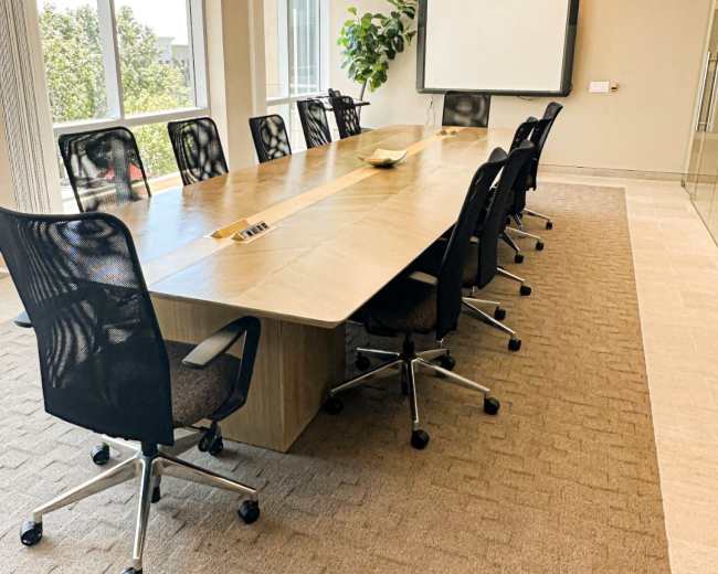 An empty conference room features a long wooden table surrounded by ergonomic chairs, with large windows allowing natural light and a presentation screen at one end.