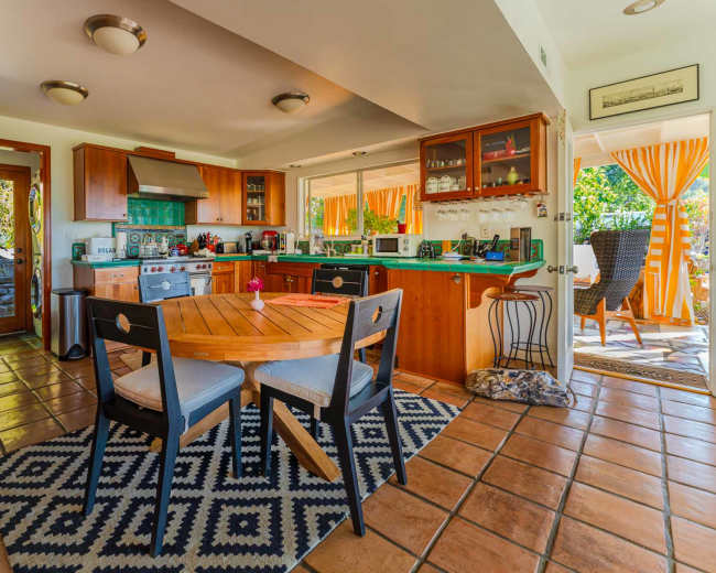 A kitchen with wooden cabinets, a round dining table, and tile flooring features large windows letting in natural light.