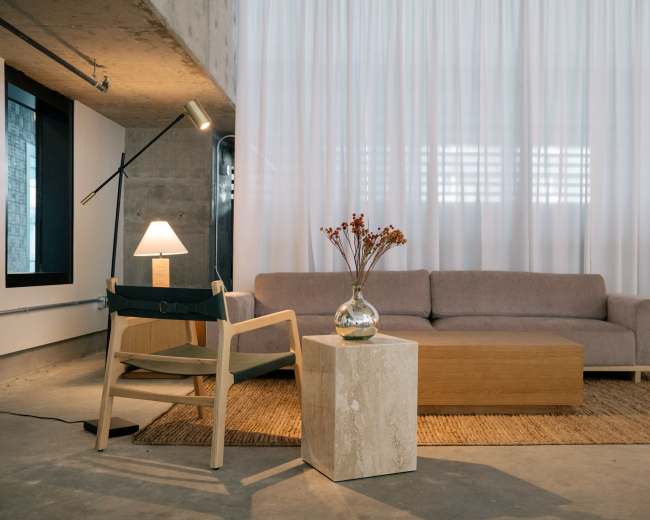 The image shows a modern living area with a sofa, a wooden coffee table, a chair, a lamp, and a vase of dried flowers against a backdrop of sheer curtains.