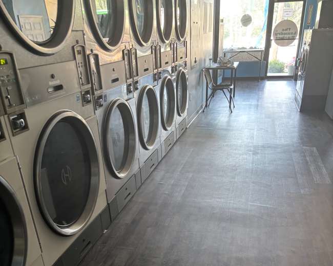 The image shows the interior of a laundromat with multiple washing machines aligned against the wall and a folding table in the middle of the room.