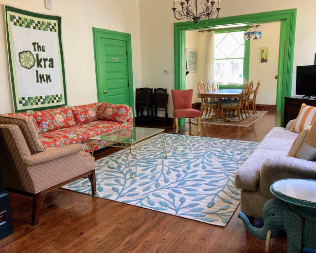 The image shows a brightly colored living room with two patterned sofas, a glass coffee table, a decorative rug, and a dining area visible through an open doorway.