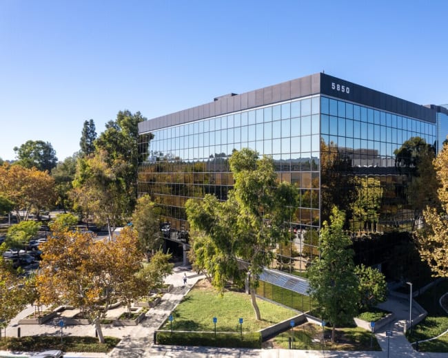 The image shows a modern glass office building surrounded by trees and a landscaped area.