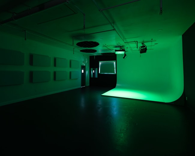 A dimly lit studio space with a green backdrop, sound-absorption panels on the walls, and overhead lighting.