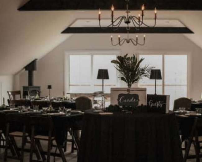 A dining area features neatly arranged tables with dark tablecloths, chairs, and a centerpiece under a chandelier.