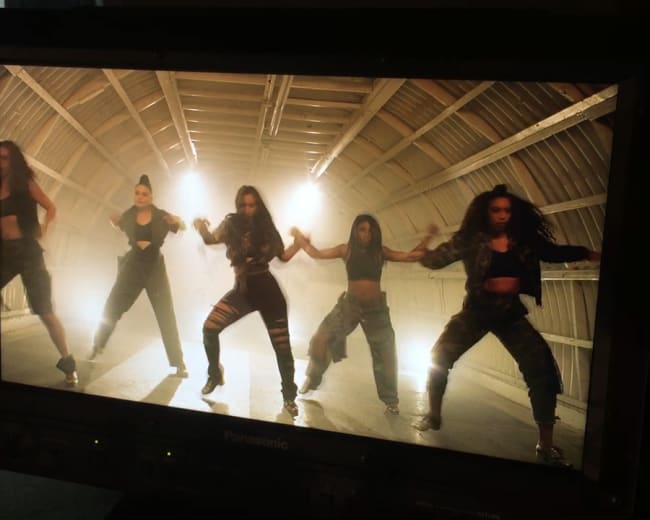 A group of five dancers performs synchronized movements in a dimly lit studio with a wooden ceiling.