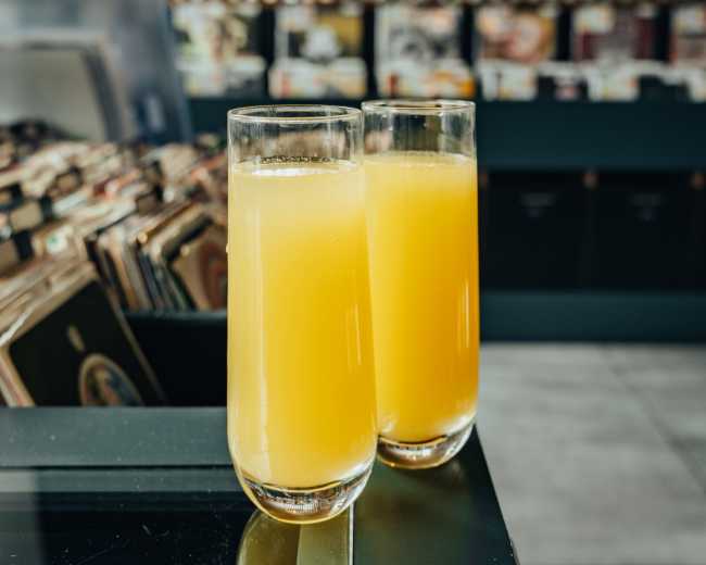 Two glasses of orange juice stand on a dark countertop in a cafe, with records visible in the background.