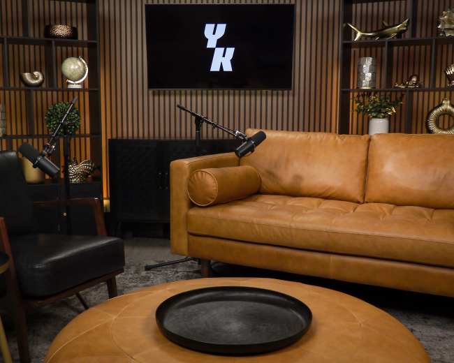 The image shows a modern chat or interview set featuring a brown leather sofa, a black armchair, a round coffee table, and a backdrop with a logo on a screen surrounded by decorative shelves.