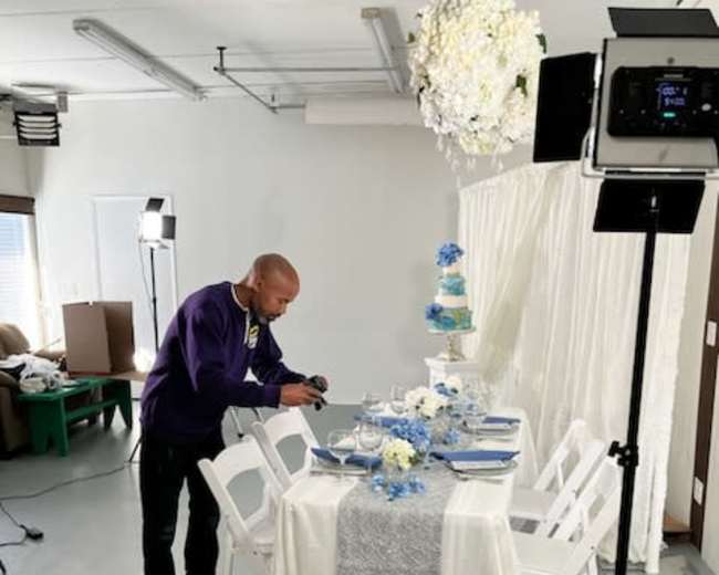 A man in a purple sweatshirt is photographing a decorated dining table set with white and blue tableware in a studio-like space.