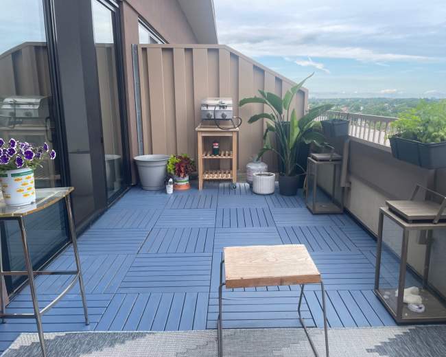 The image shows a balcony featuring outdoor furniture, potted plants, and a small grill, with a view of the sky in the background.