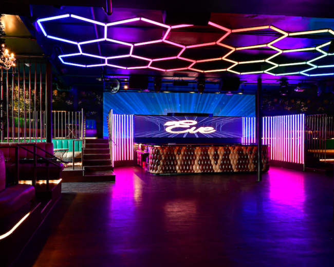 The image shows a vibrant nightclub interior featuring a geometric, illuminated ceiling and a bar area with colored lighting.