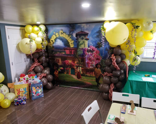 A decorated party room featuring a vibrant backdrop with a tropical theme, flanked by colorful balloons and gift bags.
