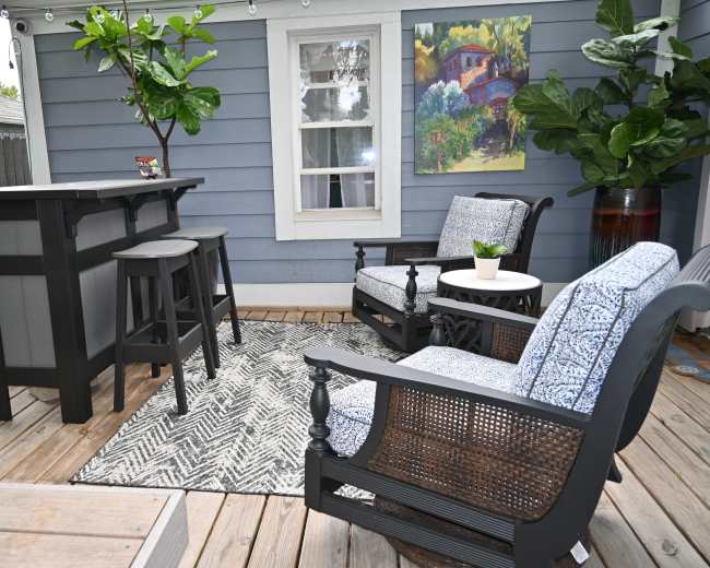 The image shows a cozy outdoor seating area with a bar table, two chairs, and a small round table on a wooden deck, surrounded by potted plants and a wall art piece.