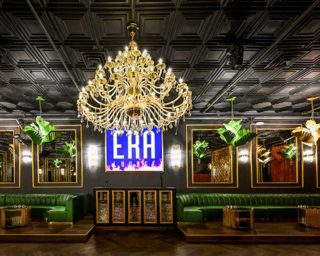 The image depicts a stylish lounge interior featuring a grand chandelier, green seating, mirrored walls, and a large illuminated sign reading "ERA."