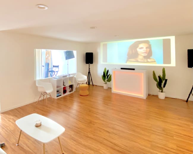 A spacious room with light hardwood flooring features a large projection screen displaying an image, surrounded by white furniture and potted plants.