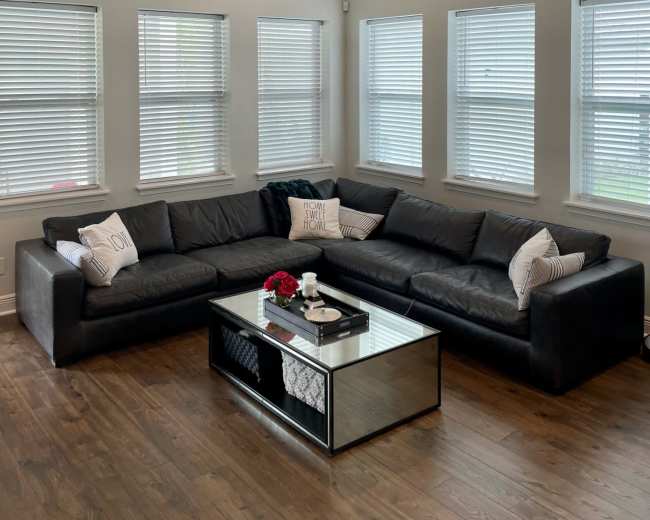 The image shows a modern living room with a large black sectional couch, a glass coffee table, and multiple windows letting in natural light.