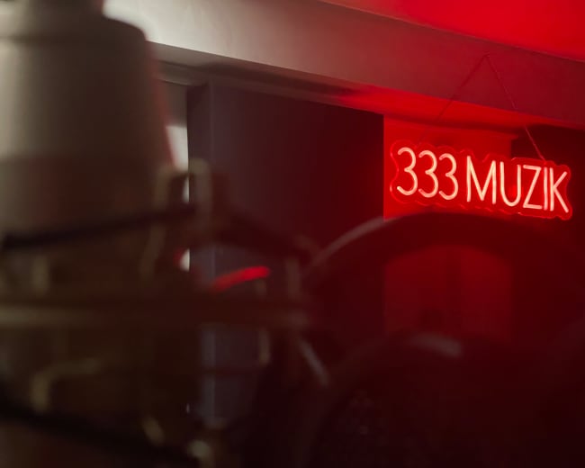 A red neon sign reading "333 MUZIK" is illuminated in a dimly lit room, with a microphone in the foreground.