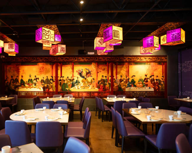 The image shows a restaurant interior with purple chairs, tables set for dining, and colorful lanterns hanging from the ceiling above a decorative mural.
