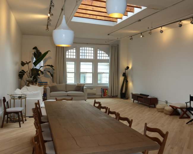 The image shows a spacious, well-lit modern dining area with a long wooden table surrounded by chairs, leading to a cozy living space that features a sofa and large windows.