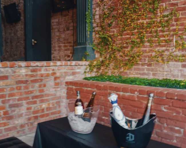 The scene features a table with two ice buckets holding bottles of champagne and whiskey against a backdrop of a brick wall with greenery.