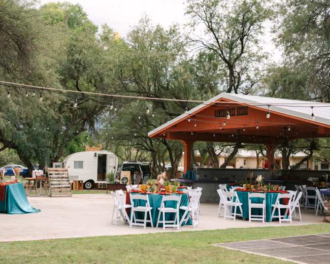 The outdoor venue features round tables with teal tablecloths set up under a covered area, surrounded by greenery and string lights, with a vintage trailer visible in the background.