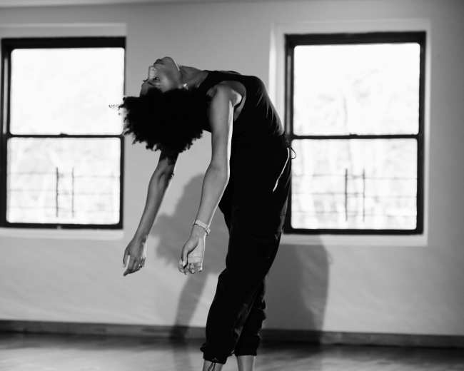 A dancer in a black outfit leans backward with arms extended in a sunlit studio with wooden floors and large windows.