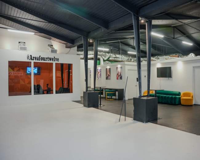 The image shows a spacious, industrial-style room with a white floor, black beams, and various seating areas, including green and yellow sofas.