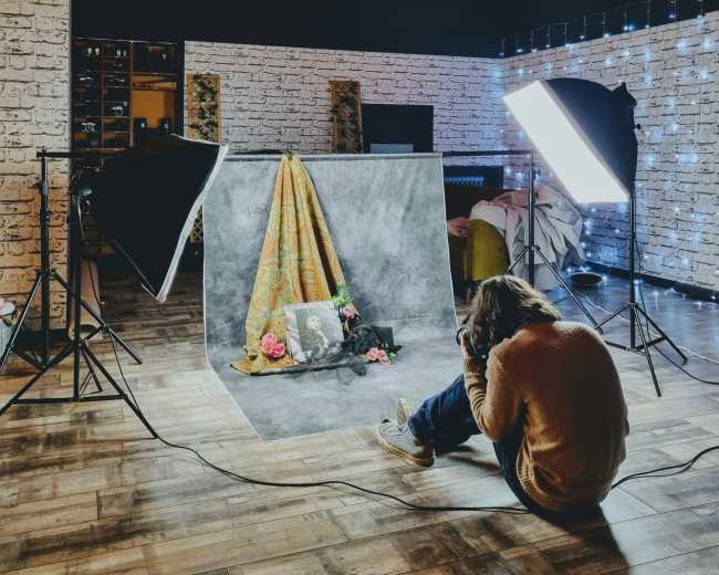 A photographer kneels in a studio, capturing an image of a decorated backdrop featuring fabric and flowers, with studio lights positioned to illuminate the scene.