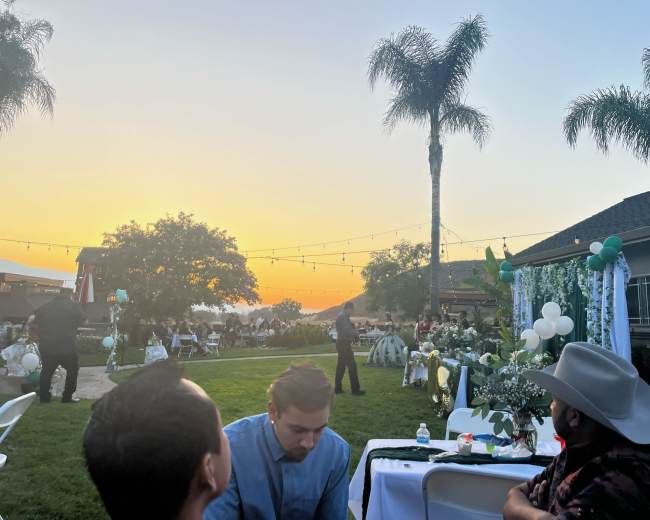 A gathering takes place outdoors at sunset, with chairs arranged around tables and decorations in a garden setting.