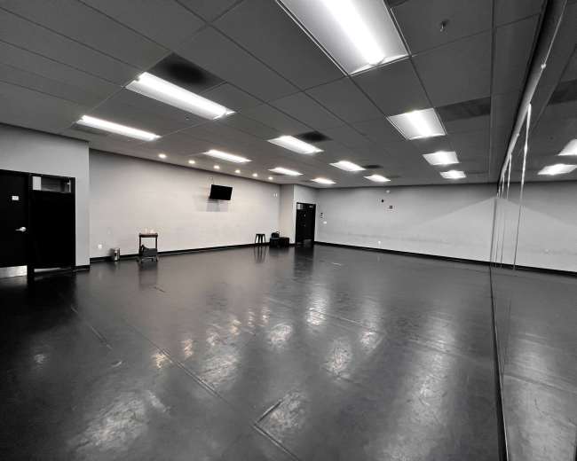 The image shows a large, empty studio space with black flooring, white walls, and a wall of mirrors.
