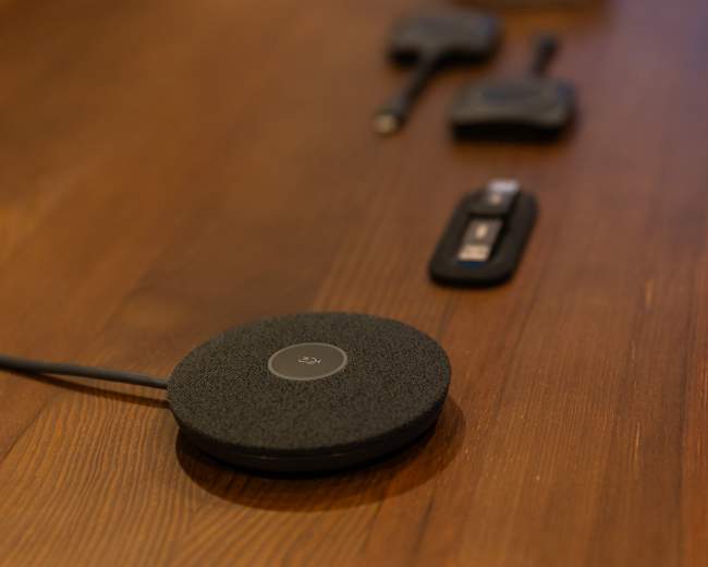 The image shows a wooden table with a row of various electronic devices, including a round black speaker, a remote control, and other small gadgets.