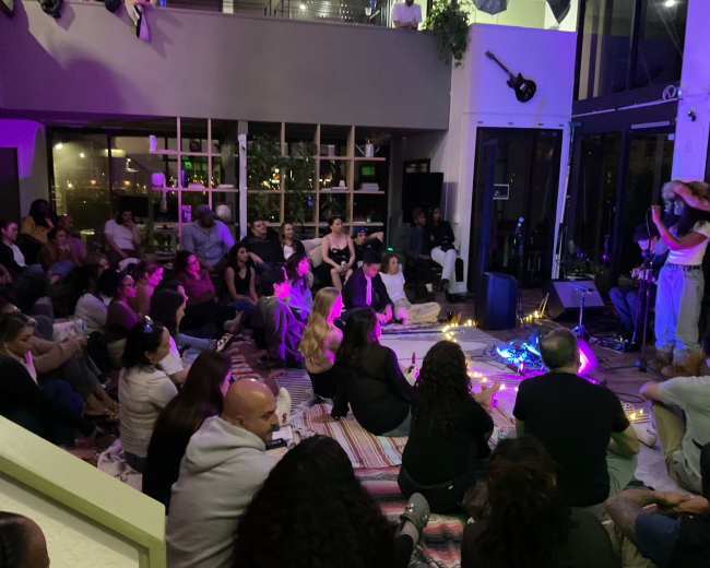 A diverse group of people sits on the floor in a cozy indoor space, attentively watching a performer with musical instruments.