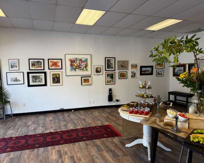 The image shows an art gallery interior with framed artwork on the walls, a table adorned with fruit and pastries, and a decorative rug on the wooden floor.