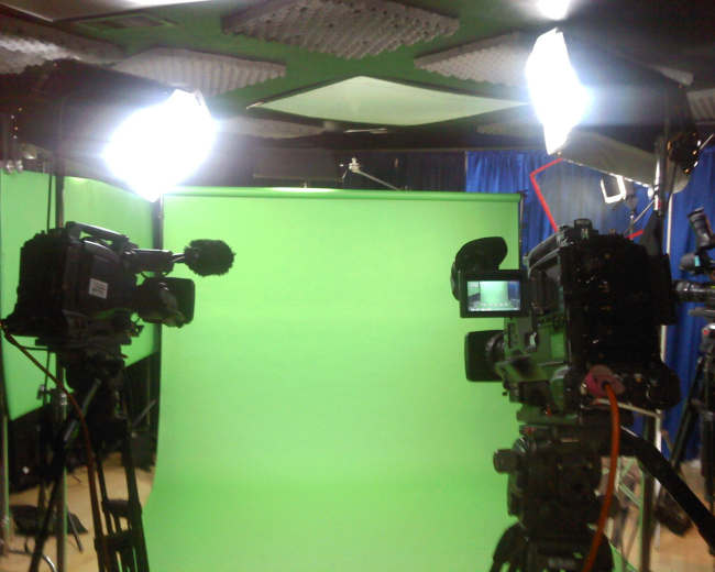 Two video cameras positioned in front of a green screen, with studio lighting set up around them.