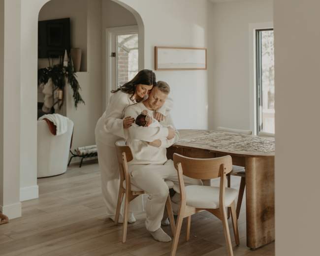 A woman embraces a child at a wooden dining table in a softly lit, modern dining area.