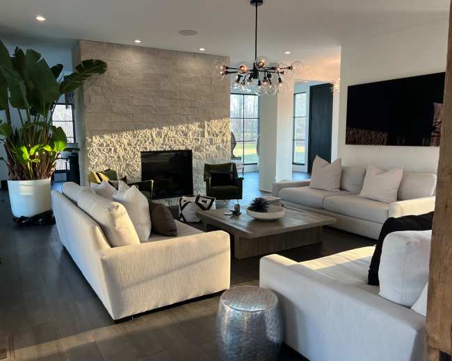 The image shows a modern living room featuring light-colored sofas, a stone fireplace, a large plant, and a coffee table in front of a window with natural light.