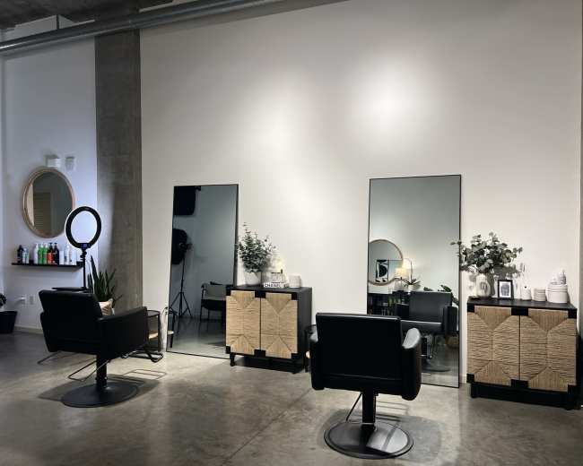The image shows a modern hair salon featuring two styling stations with mirrors, black chairs, and decorative elements on wooden cabinets against a neutral wall.
