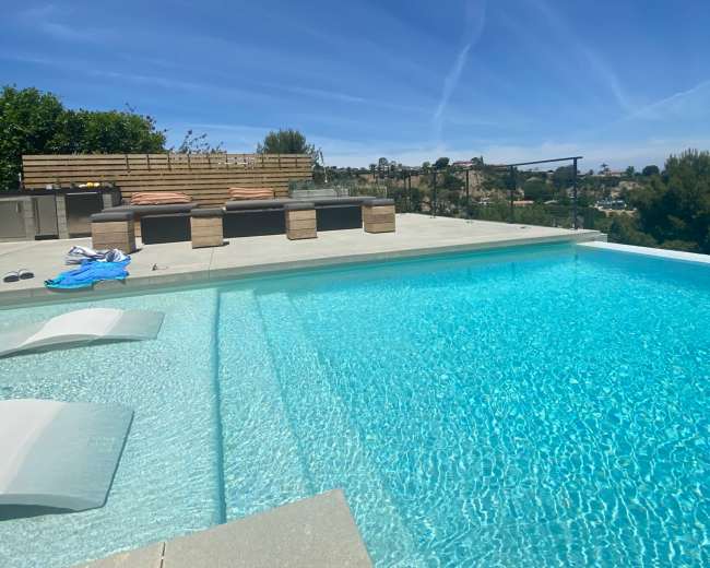 The image shows a clear swimming pool with a sunbathing area and lounge chairs against a backdrop of blue skies and distant hills.