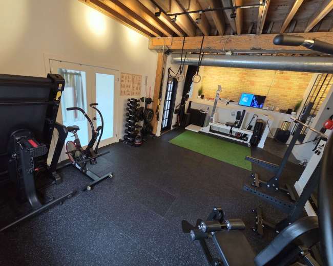 The image shows a well-equipped home gym featuring cardio and weight training equipment on black rubber flooring.