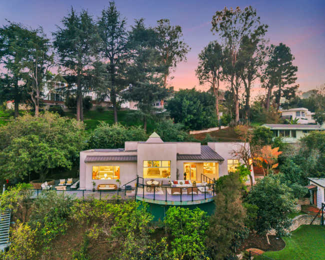 A modern house with large windows is situated on a hillside, surrounded by trees and greenery.